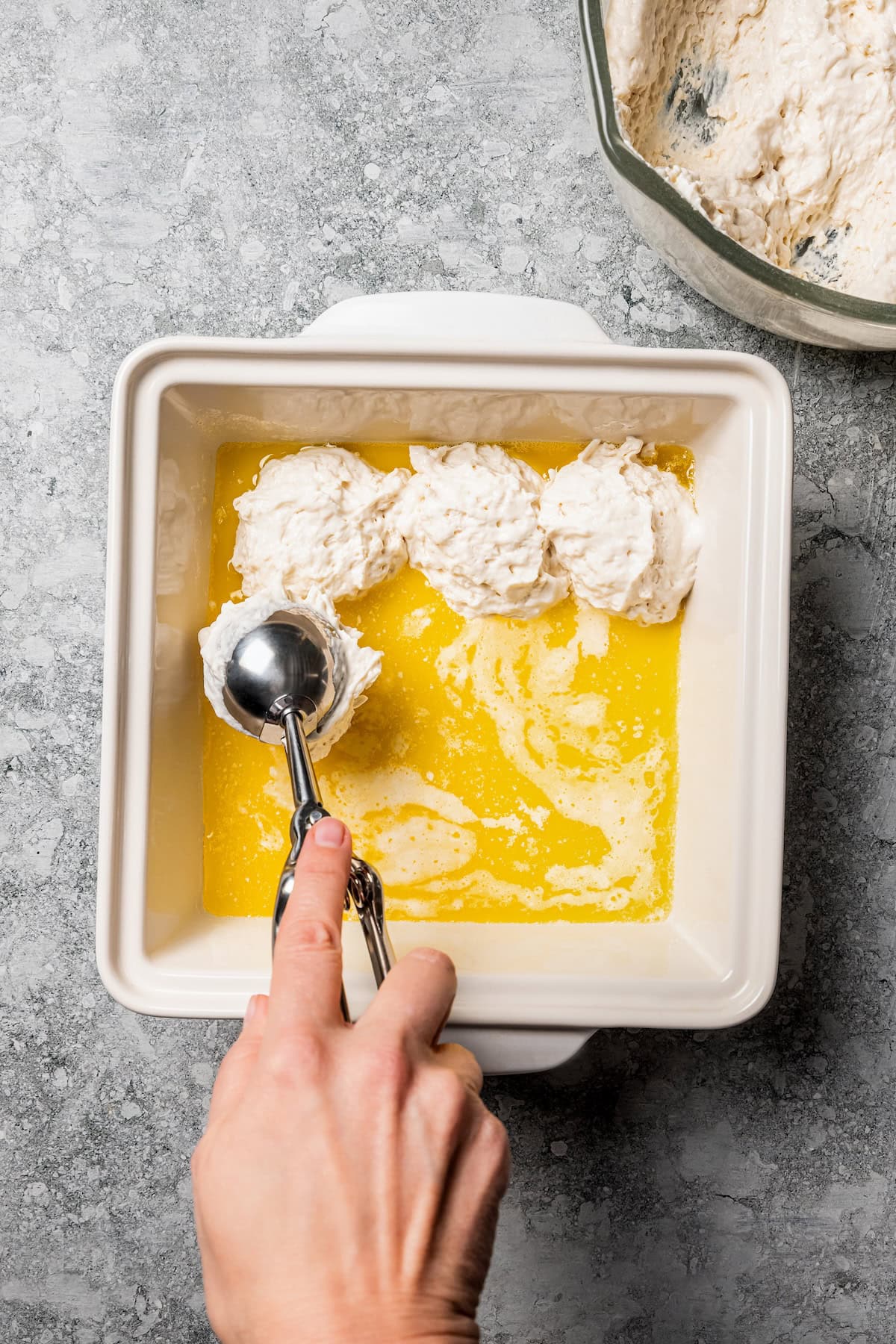 Butter swim biscuits are fluffy drop biscuits baked in a pool of butter! Crispy outside, soft inside, and easy to make with 5 ingredients. A hand using a large cookie scoop to place scoops of biscuit batter on top of a layer of melted butter in a square baking dish.