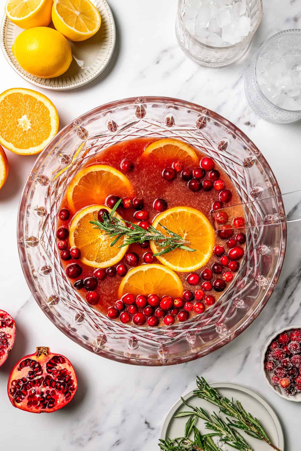 This festive Christmas punch recipe is the best big-batch cocktail to get your holiday party started this year. Make it boozy, or not! Overhead view of homemade Christmas punch in a punch bowl garnished with orange slices, cranberries, and a rosemary sprig, surrounded by punch ingredients.