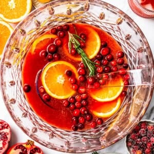 Christmas punch in a bowl, and it's garnished with orange slices, cranberries, and a rosemary sprig, surrounded by punch ingredients.