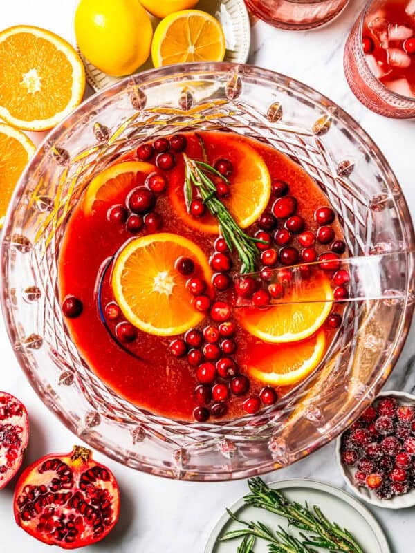 Christmas punch in a bowl, and it's garnished with orange slices, cranberries, and a rosemary sprig, surrounded by punch ingredients.