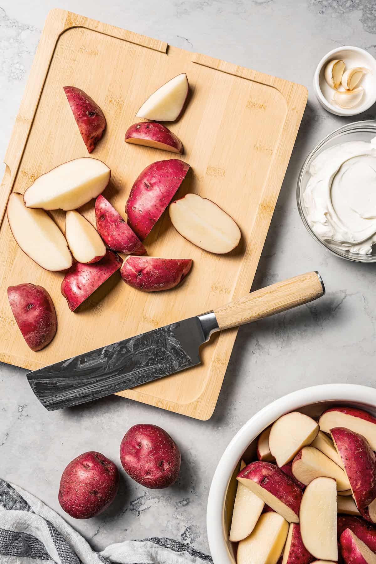 These slow cooker garlic mashed potatoes are made with creamy, skin-on red potatoes and plenty of butter! No boiling, and only 6 ingredients. Overhead view of a knife resting next to chopped red potatoes on a wooden cutting board.