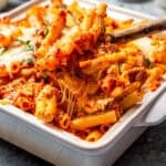 A spoon scooping cheesy baked ziti from a baking dish.