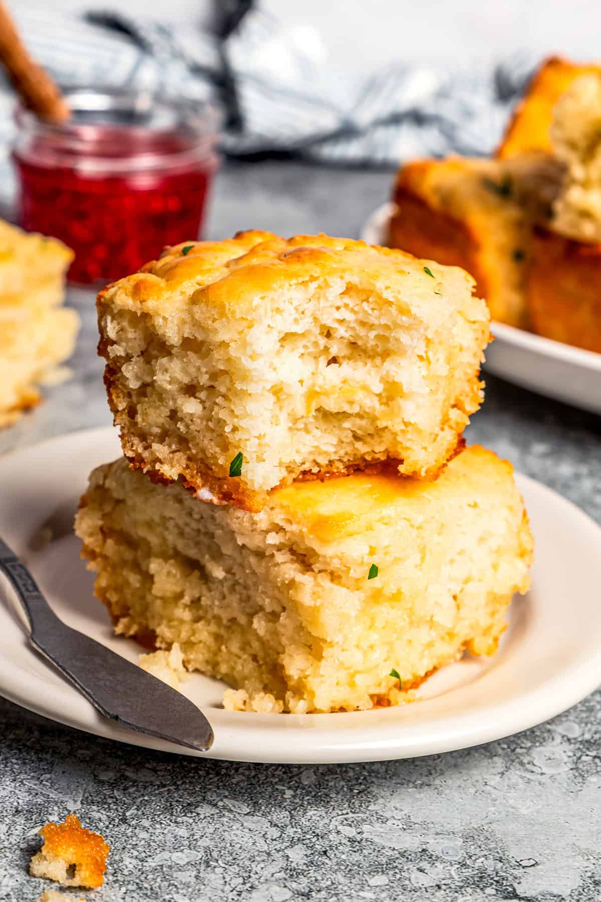 Butter swim biscuits are fluffy drop biscuits baked in a pool of butter! Crispy outside, soft inside, and easy to make with 5 ingredients. Two butter swim biscuits stacked on a plate with a bite missing from the top biscuit, with jar of jam and more biscuits in the background.