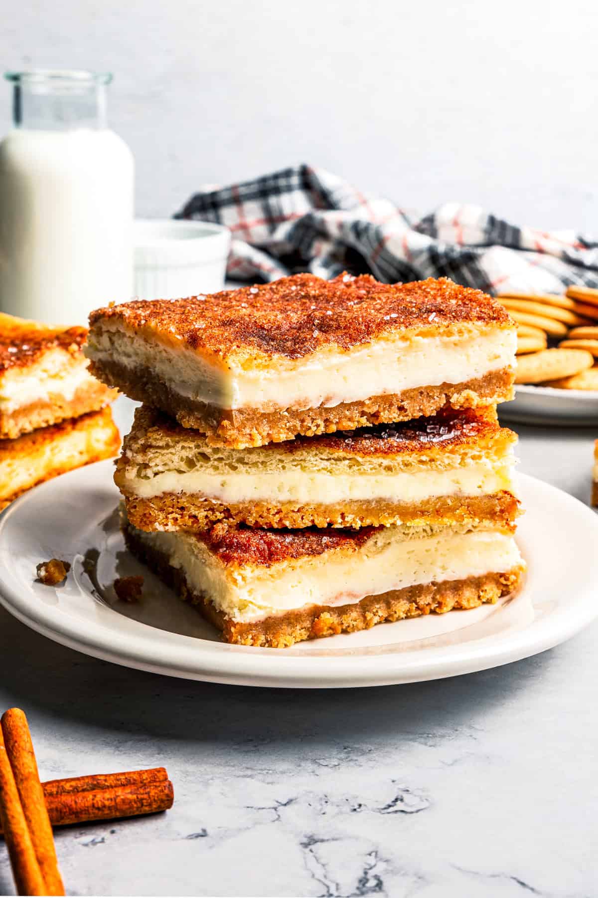 This churro cheesecake bars recipe is an easy dessert with layers of cinnamon sugar, a flaky pastry crust, and creamy cheesecake filling. Three churro cheesecake bars stacked on a white plate with a jug of milk and more cheesecake bars in the background.