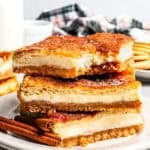 Churro cheesecake bars stacked on a white plate with a jug of milk and more cheesecake bars in the background.