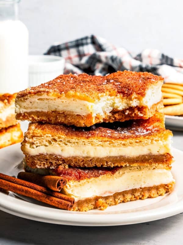 Churro cheesecake bars stacked on a white plate with a jug of milk and more cheesecake bars in the background.