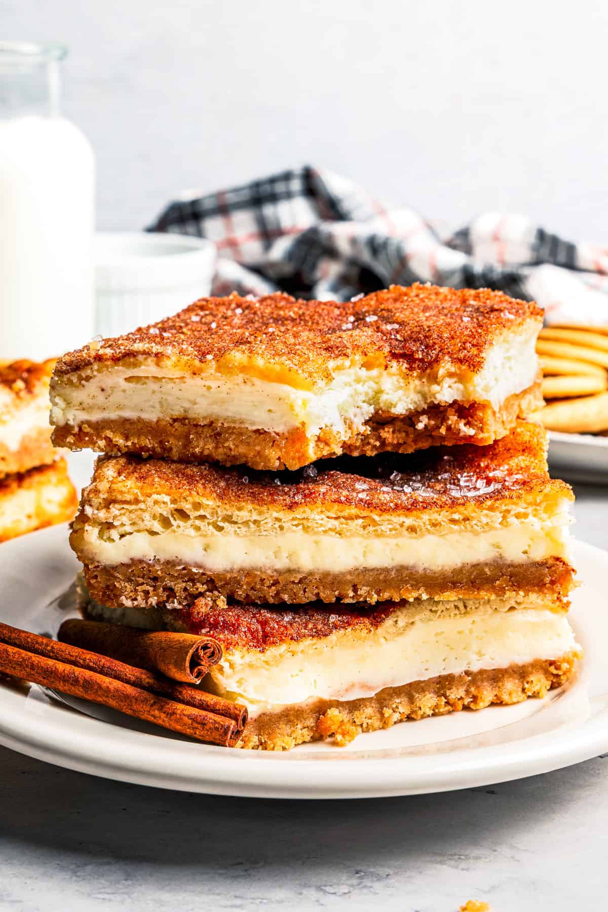 This churro cheesecake bars recipe is an easy dessert with layers of cinnamon sugar, a flaky pastry crust, and creamy cheesecake filling. Churro cheesecake bars stacked on a white plate with a jug of milk and more cheesecake bars in the background.