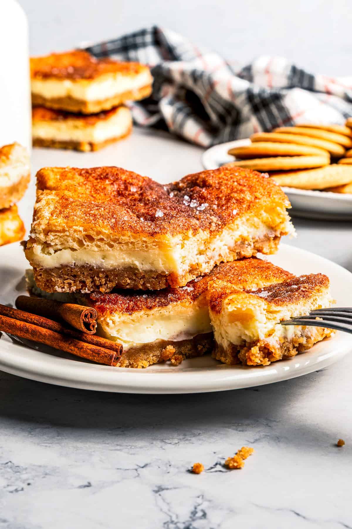 This churro cheesecake bars recipe is an easy dessert with layers of cinnamon sugar, a flaky pastry crust, and creamy cheesecake filling. Churro cheesecake bars stacked on a plate with a jug of milk and more cheesecake bars in the background.