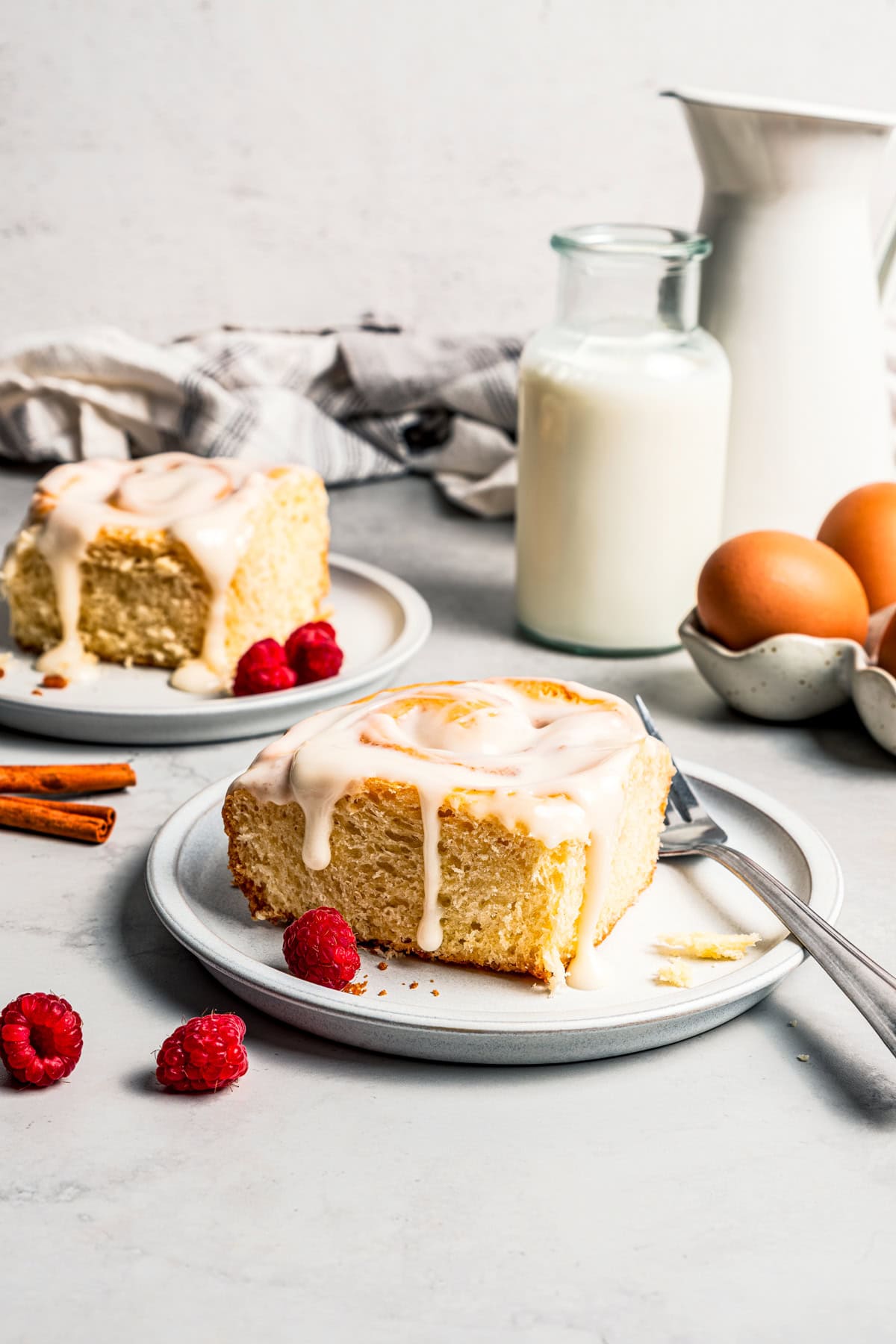 This cinnamon roll recipe makes big, bakery-style cinnamon buns filled with gooey cinnamon sugar and topped with sweet cream cheese frosting. A frosted cinnamon roll on a plate with a second cinnamon roll next to a jug of milk in the background.