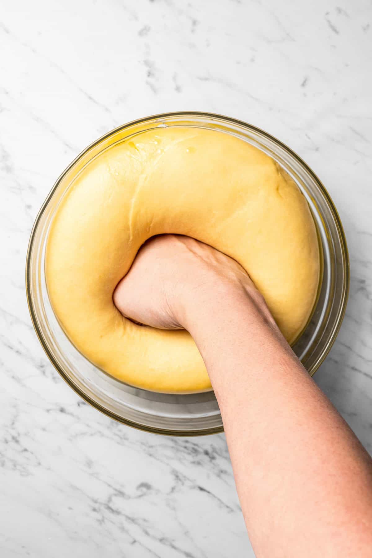 A hand punching down risen challah dough in a glass bowl.