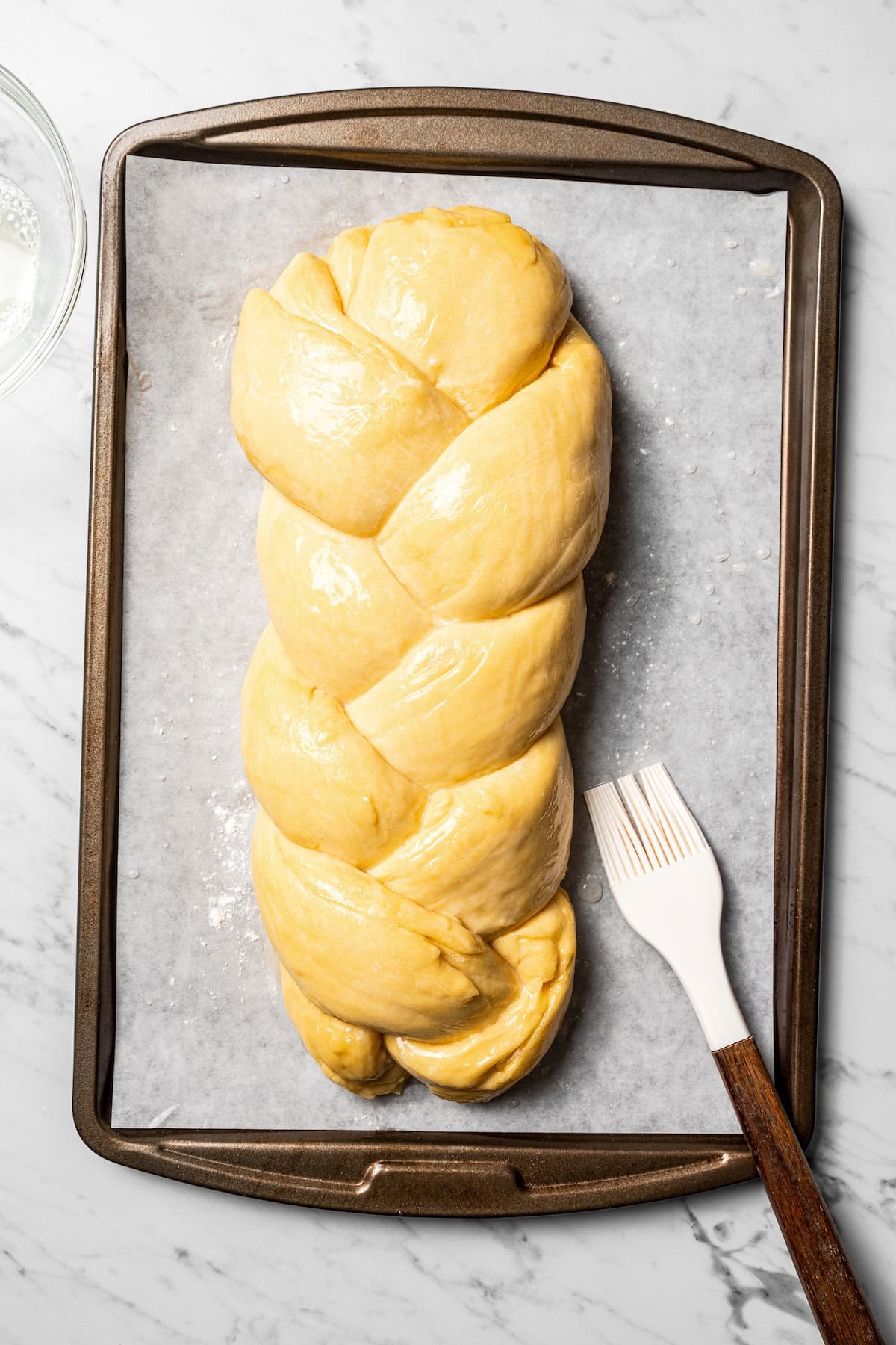 Risen challah braid brushed with an egg wash on a parchment-lined baking sheet.