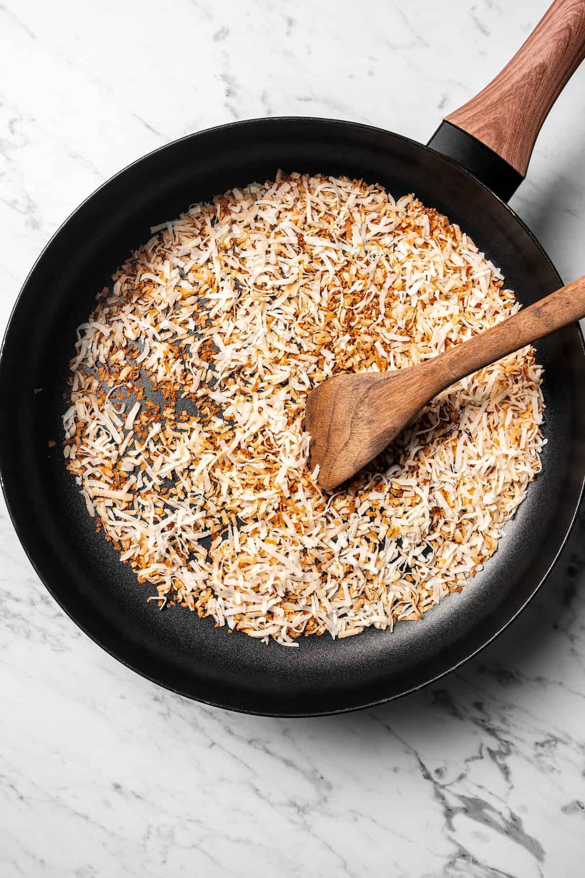 This easy coconut cream pie recipe features a thick, creamy coconut filling topped with fresh whipped cream in a golden, flaky pie crust. Coconut toasting in a skillet with a wooden spoon.