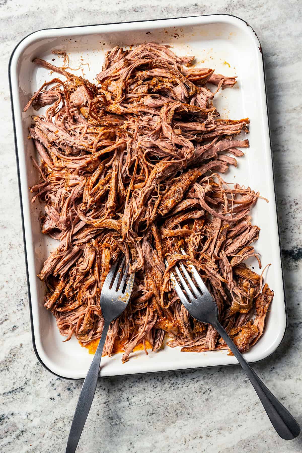 Crock pot carne asada has smoky spices and bright citrus, soaked into juicy shredded steak. Only 5 minutes to prep and perfect for tacos! Two forks resting on a tray of shredded carne asada.