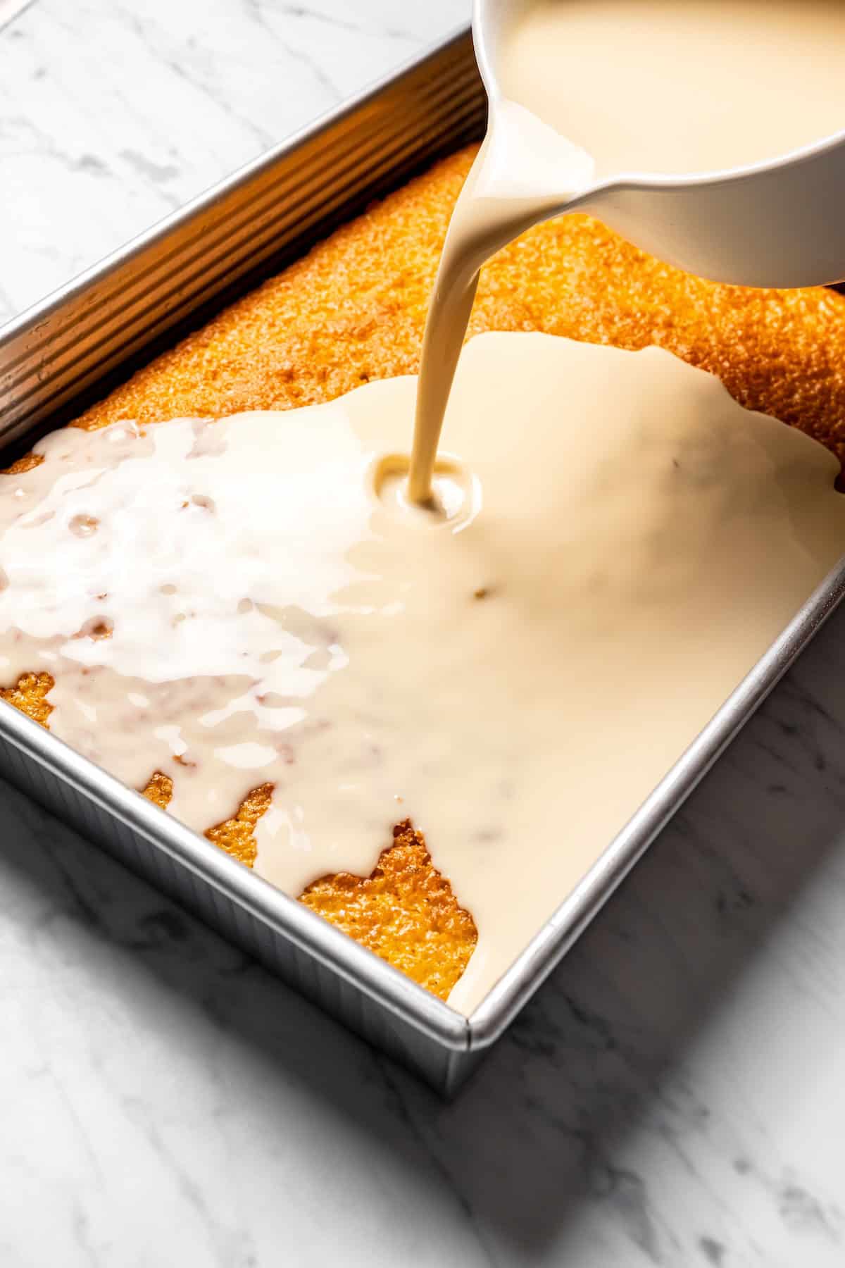 This authentic Mexican tres leches cake is a light, spongy cake soaked in a sweet milk mixture and topped with fluffy homemade whipped cream. Pouring milk sauce on top of a baked tres leches cake in a pan.
