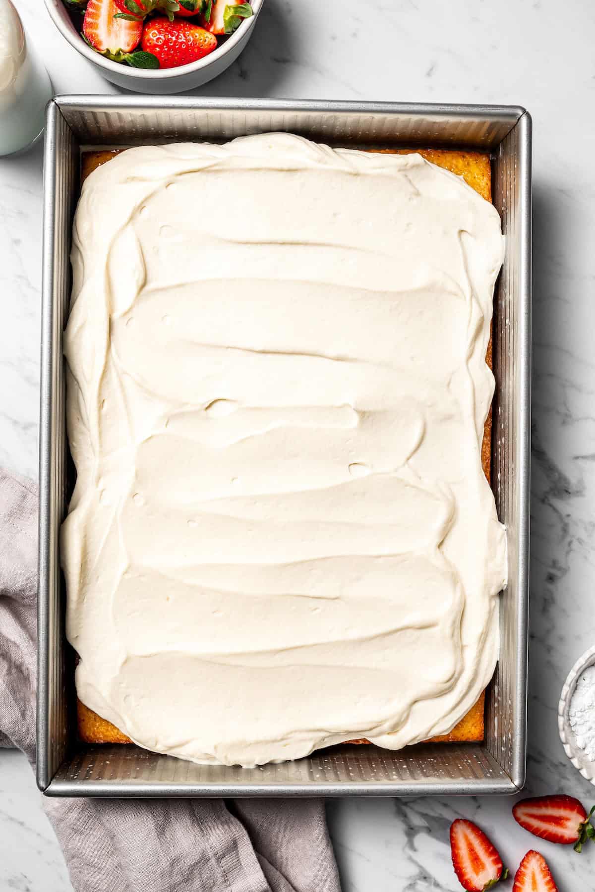 This authentic Mexican tres leches cake is a light, spongy cake soaked in a sweet milk mixture and topped with fluffy homemade whipped cream. Overhead view of tres leches cake topped with whipped cream in a baking pan.