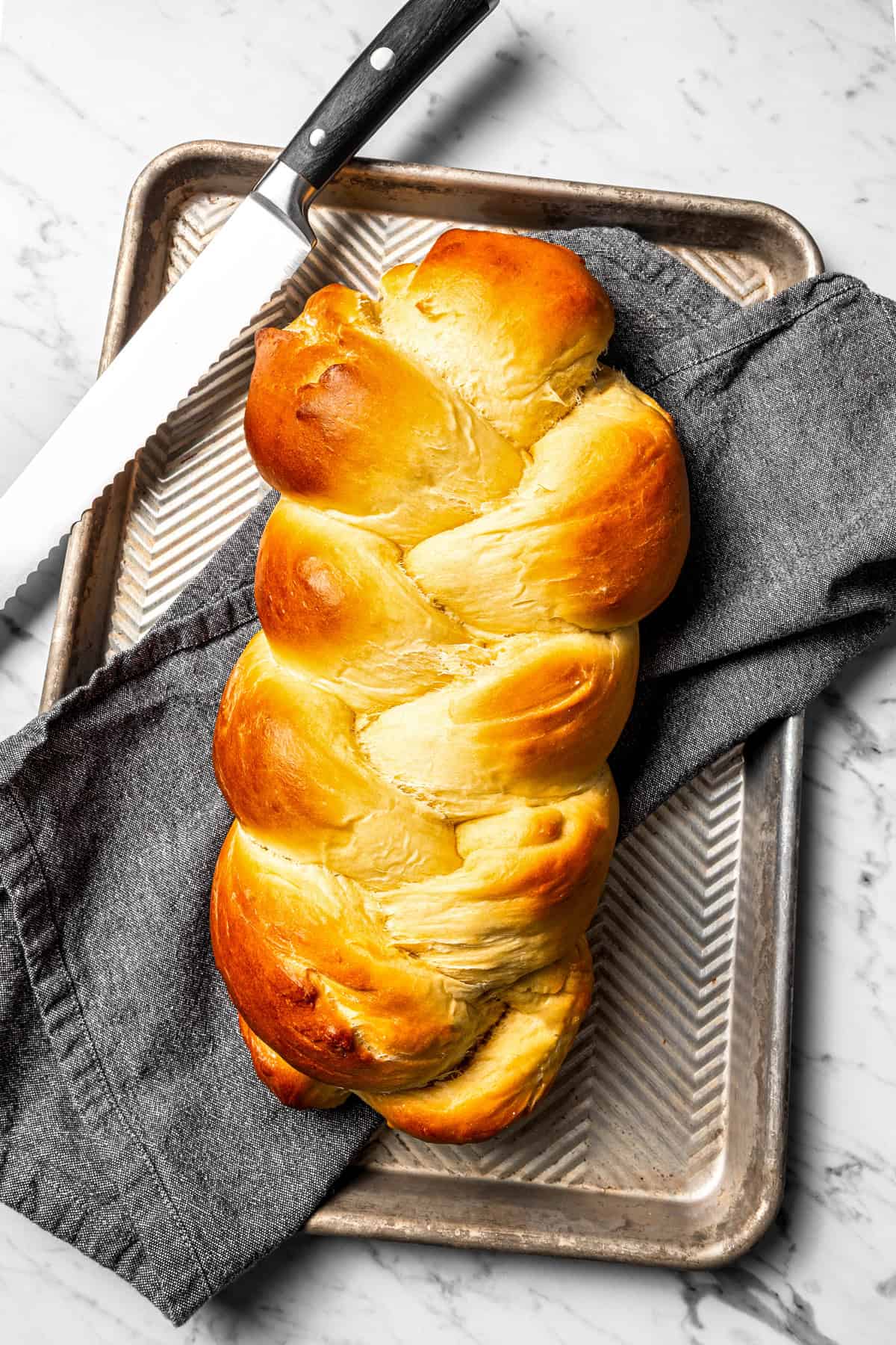 Baked challah next to a bread knife on a baking sheet lined with a dishcloth.