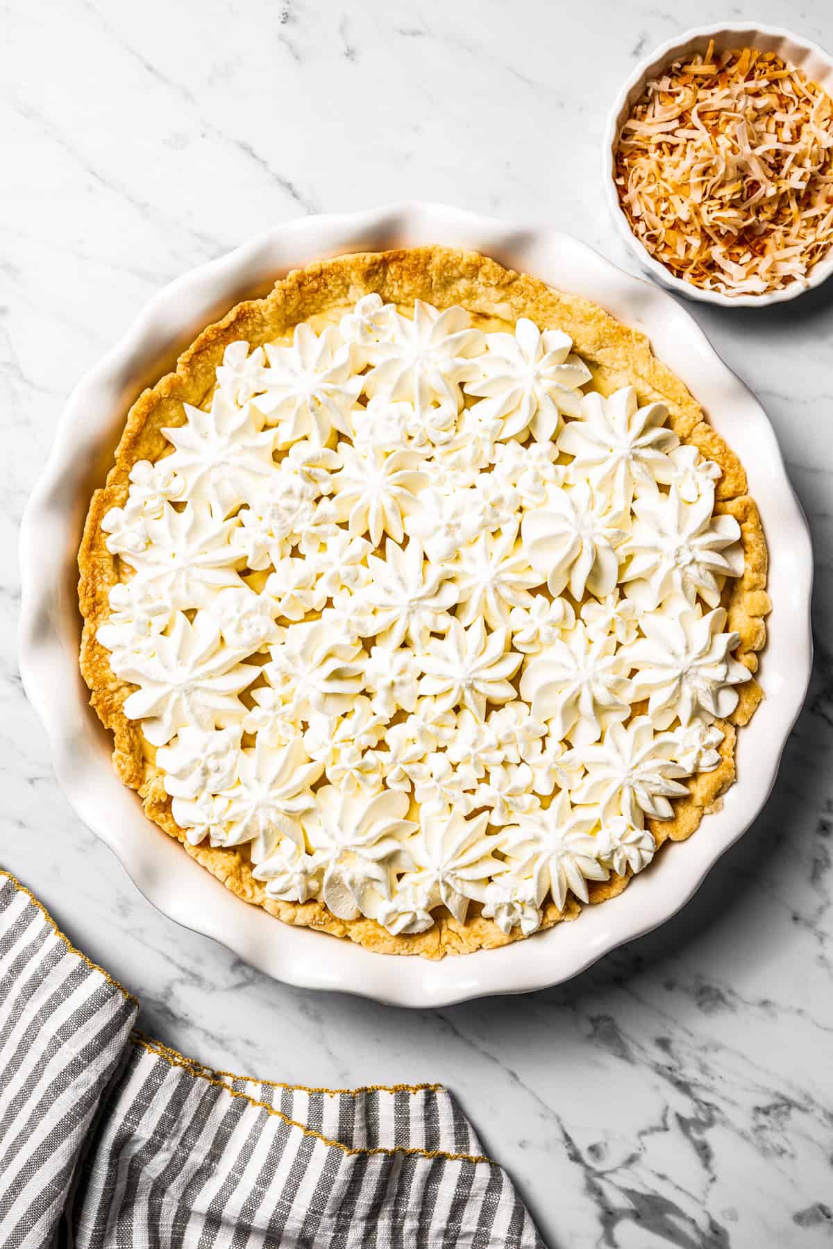 This easy coconut cream pie recipe features a thick, creamy coconut filling topped with fresh whipped cream in a golden, flaky pie crust. Overhead view of coconut cream pie topped with piped whipped cream in a pie plate.