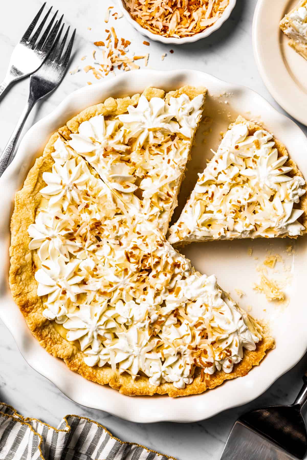 This easy coconut cream pie recipe features a thick, creamy coconut filling topped with fresh whipped cream in a golden, flaky pie crust. Coconut cream pie in a pie plate with one slice cut and a second slice missing.