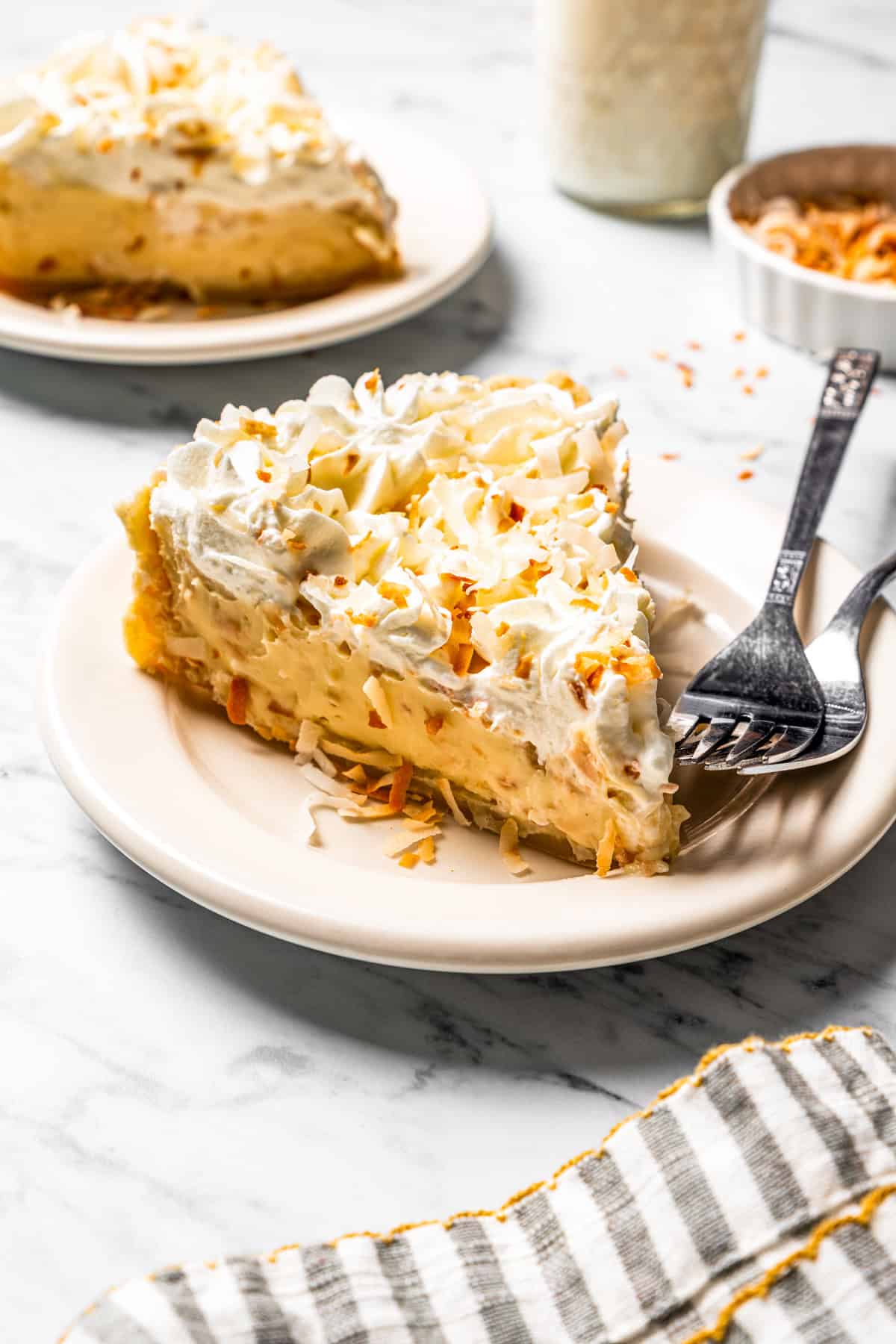 This easy coconut cream pie recipe features a thick, creamy coconut filling topped with fresh whipped cream in a golden, flaky pie crust. A slice of coconut cream pie on a plate with a second slice in the background.