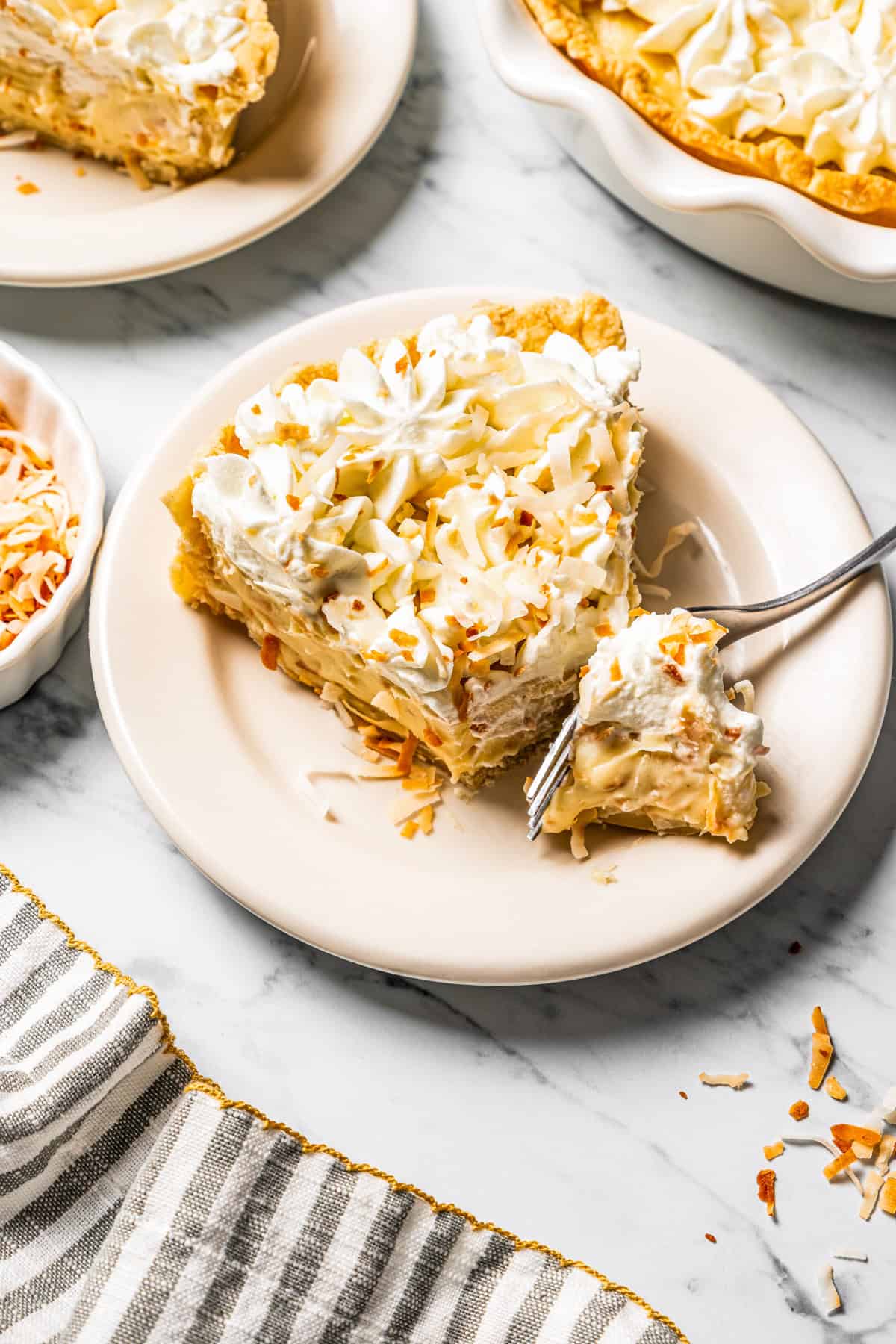 This easy coconut cream pie recipe features a thick, creamy coconut filling topped with fresh whipped cream in a golden, flaky pie crust. A fork cutting into a slice of coconut cream pie served on a plate.