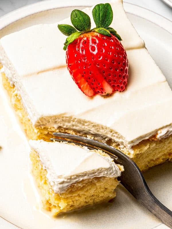 A fork cutting into the corner of a slice of tres leches cake topped with a sliced strawberry on a plate.