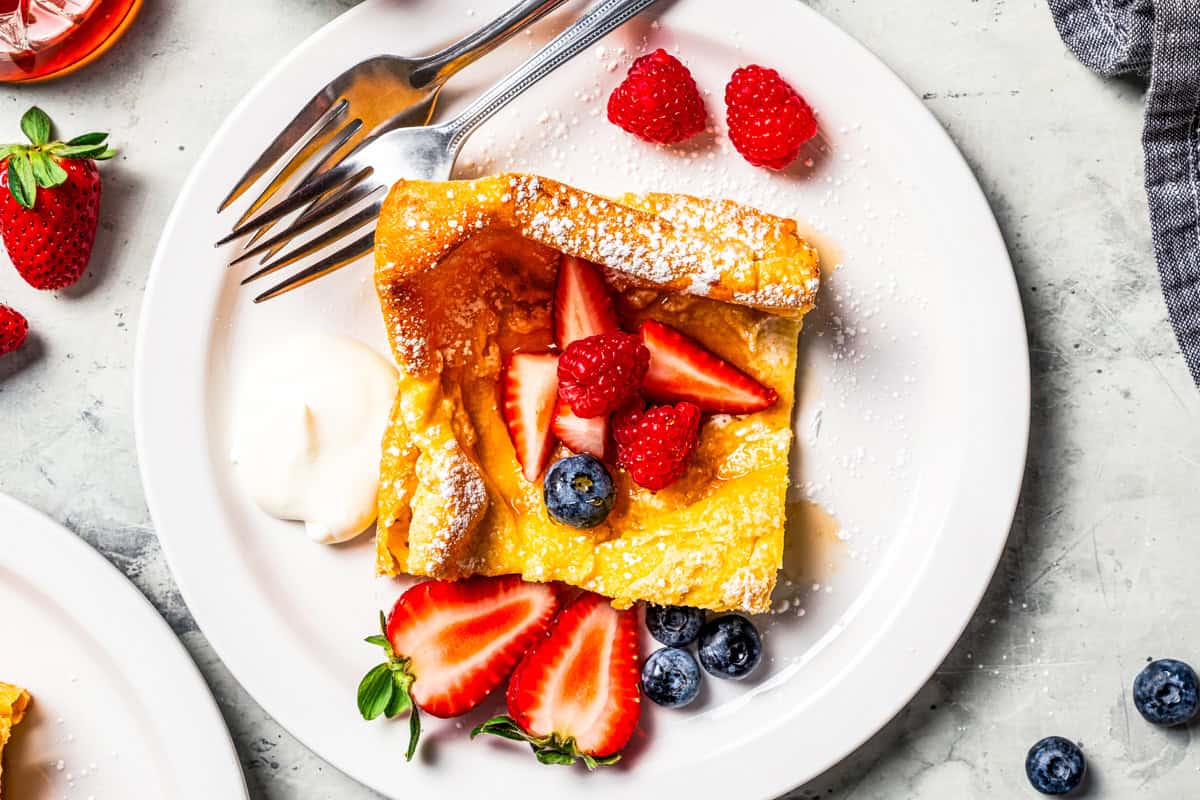 This German pancake recipe takes minutes to prepare and bakes up super soft, puffed, and golden in the oven. Made with only 6 ingredients! Overhead view of a German pancake slice garnished with fresh berries and powdered sugar on a plate with a fork.