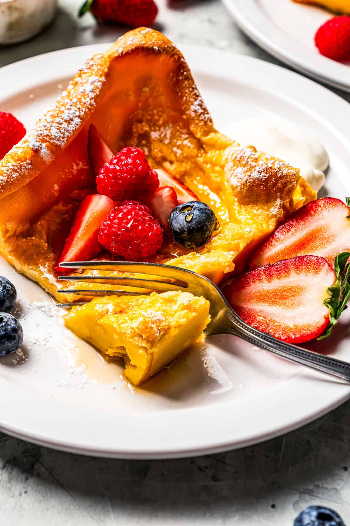 A fork cutting into the corner of a German pancake slice topped with fresh berries, next to a dollop of yogurt on a plate.
