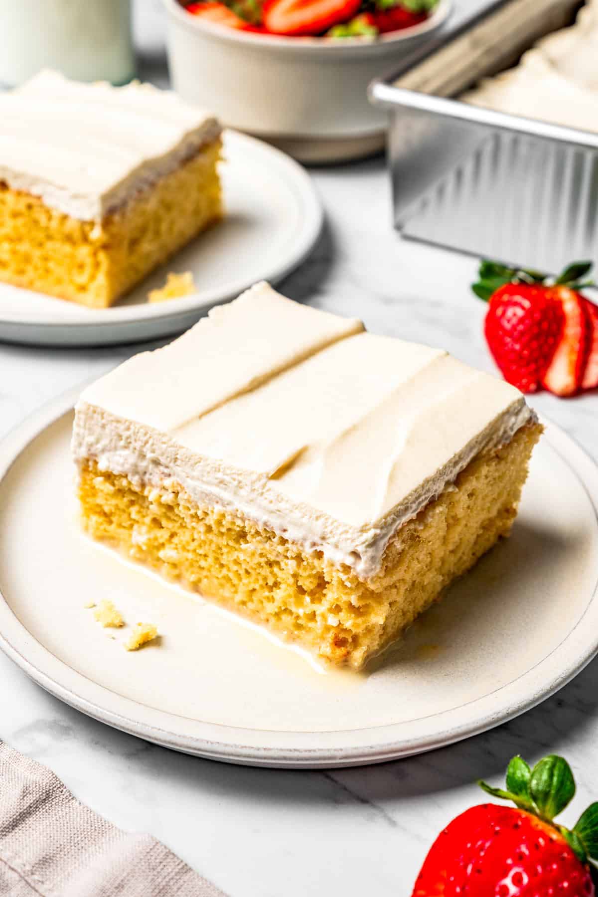 This authentic Mexican tres leches cake is a light, spongy cake soaked in a sweet milk mixture and topped with fluffy homemade whipped cream. A slice of tres leches cake on a plate with a second slice of cake in the background.
