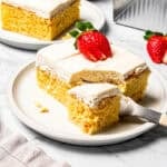 A fork cutting into the corner of a slice of tres leches cake topped with a sliced strawberry on a plate.