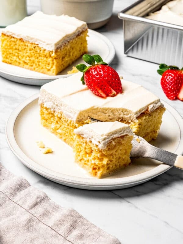A fork cutting into the corner of a slice of tres leches cake topped with a sliced strawberry on a plate.