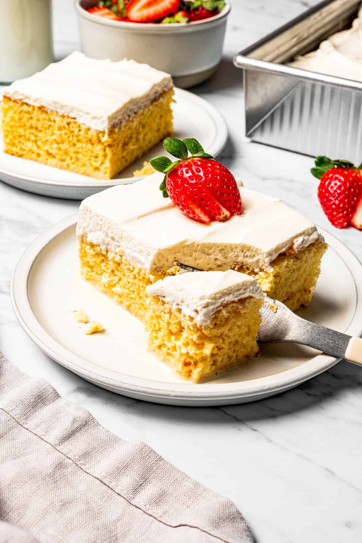 This authentic Mexican tres leches cake is a light, spongy cake soaked in a sweet milk mixture and topped with fluffy homemade whipped cream. A fork cutting into the corner of a slice of tres leches cake topped with a sliced strawberry on a plate.