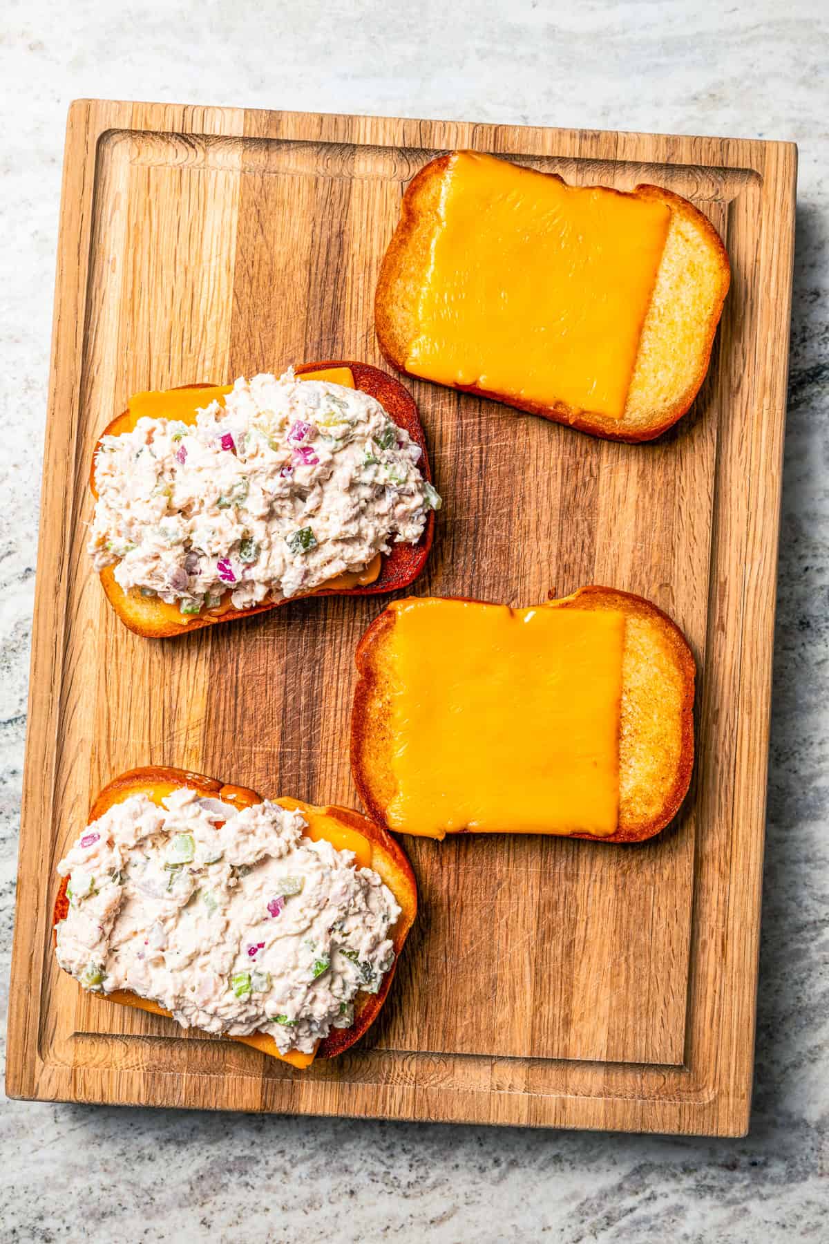 Two toasted bread slices topped with melted cheese, and two slices topped with tuna salad on a cutting board.
