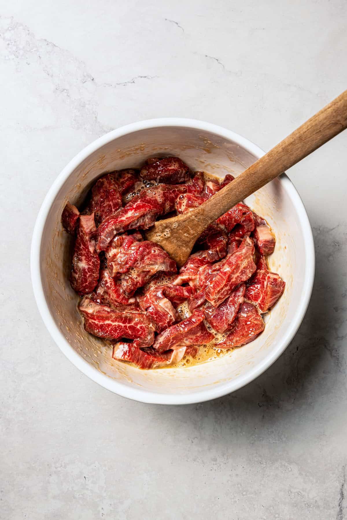 Beef strips marinating in a bowl with a spatula.