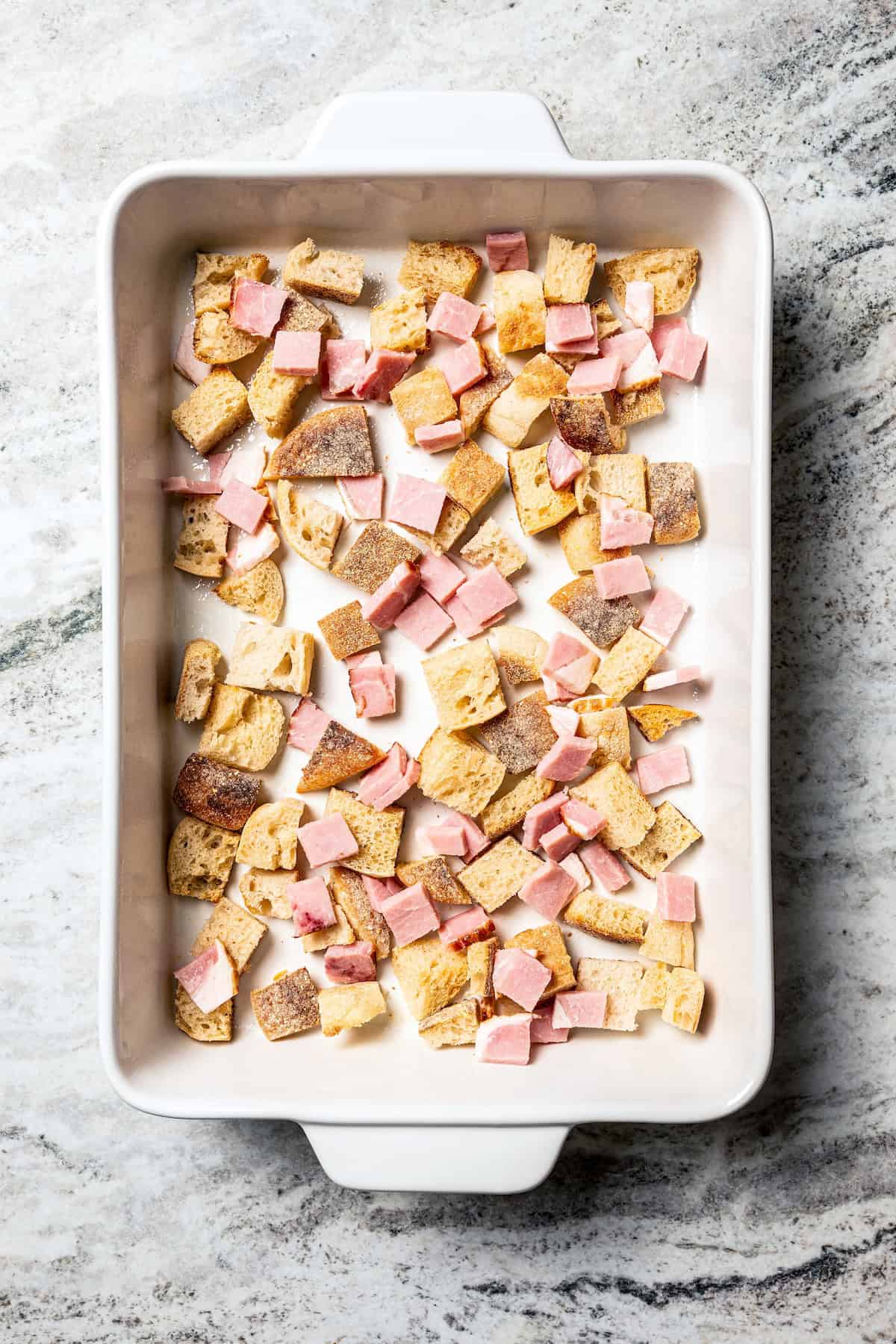The first layer of cubed English muffins and chopped Canadian bacon in the bottom of a baking dish.