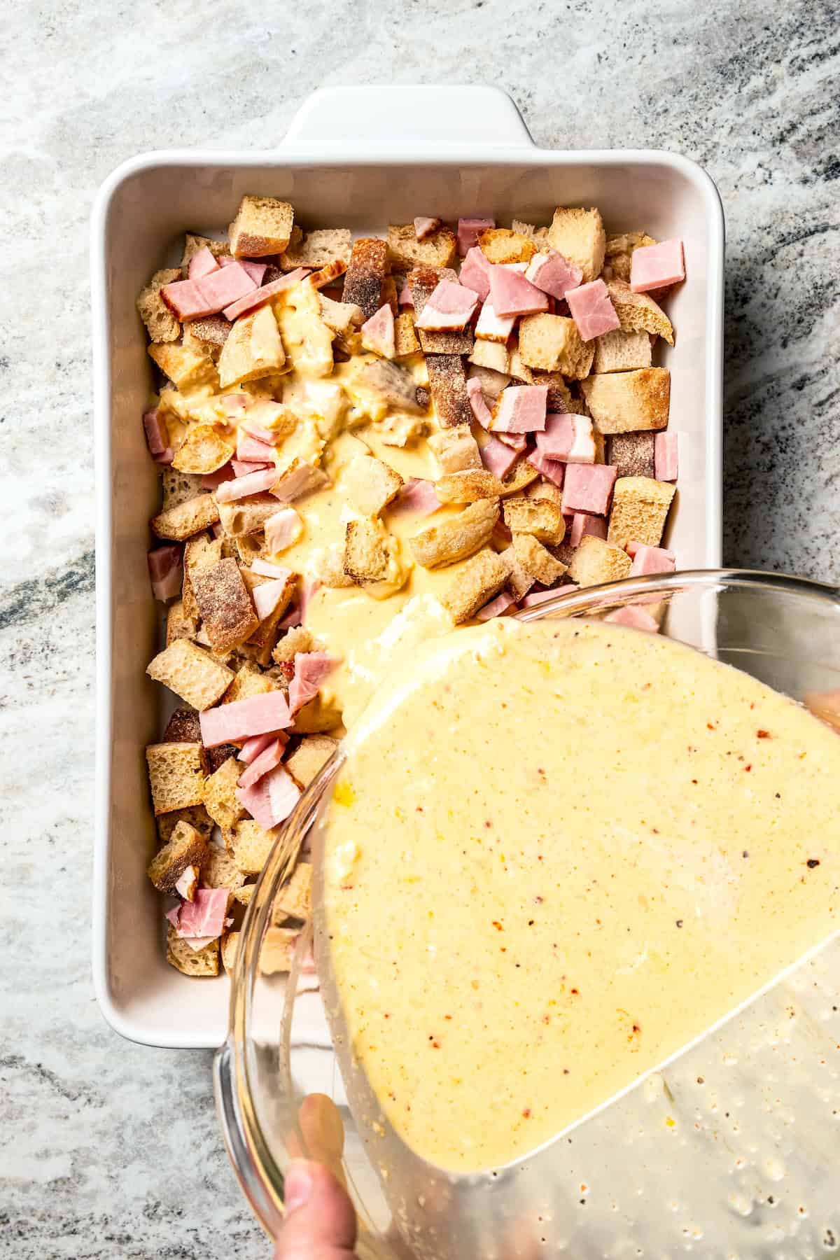 Custard being poured over cubed English muffins and bacon in a baking dish.