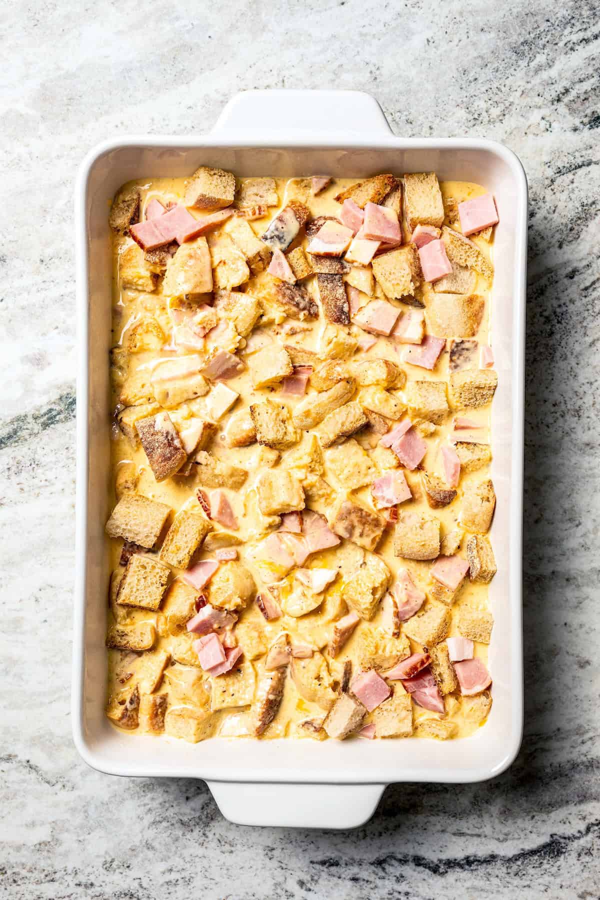 English muffins and bacon soaked with custard in a baking dish.