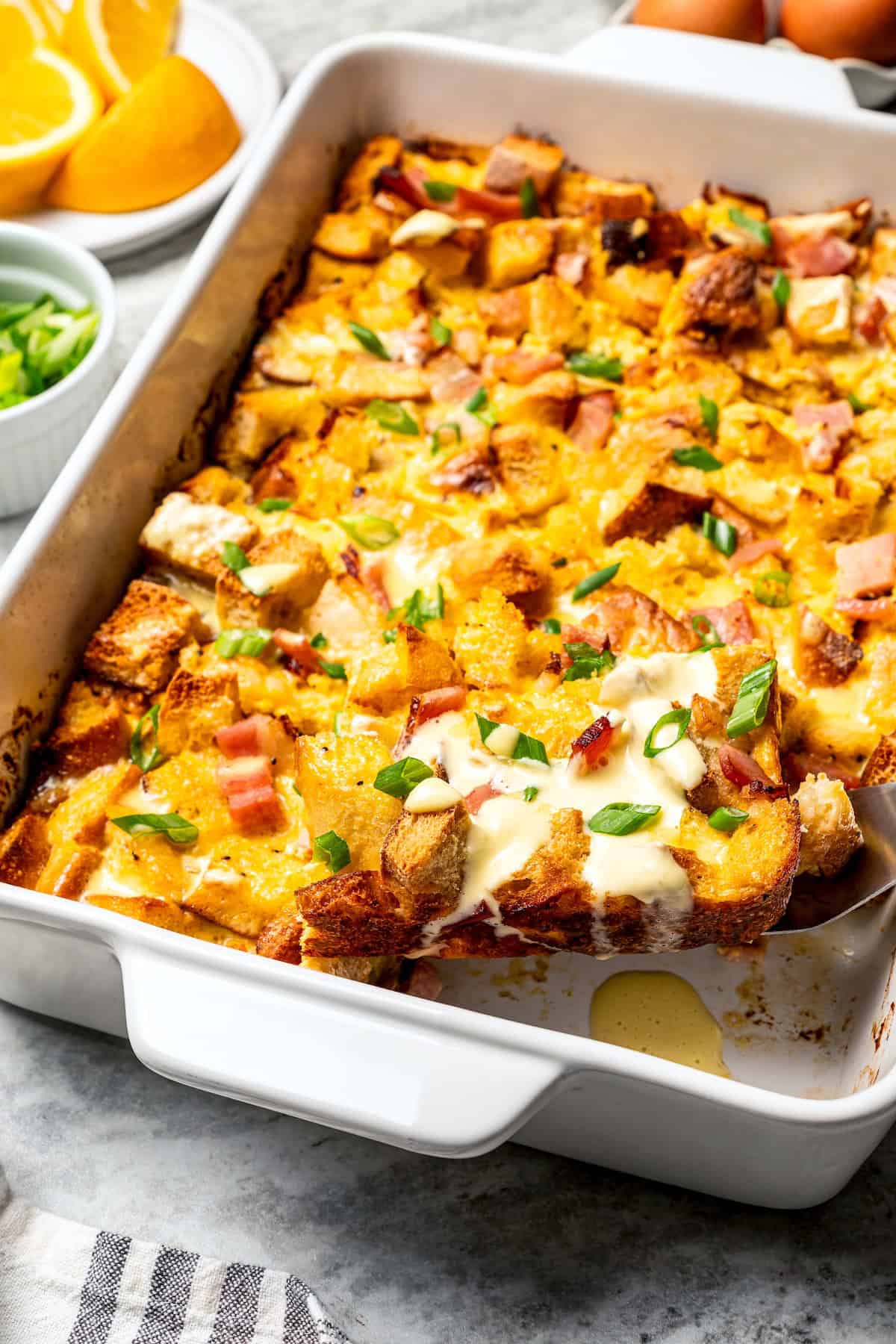 A slice of Eggs Benedict casserole being lifted from the rest of the casserole in a baking dish.