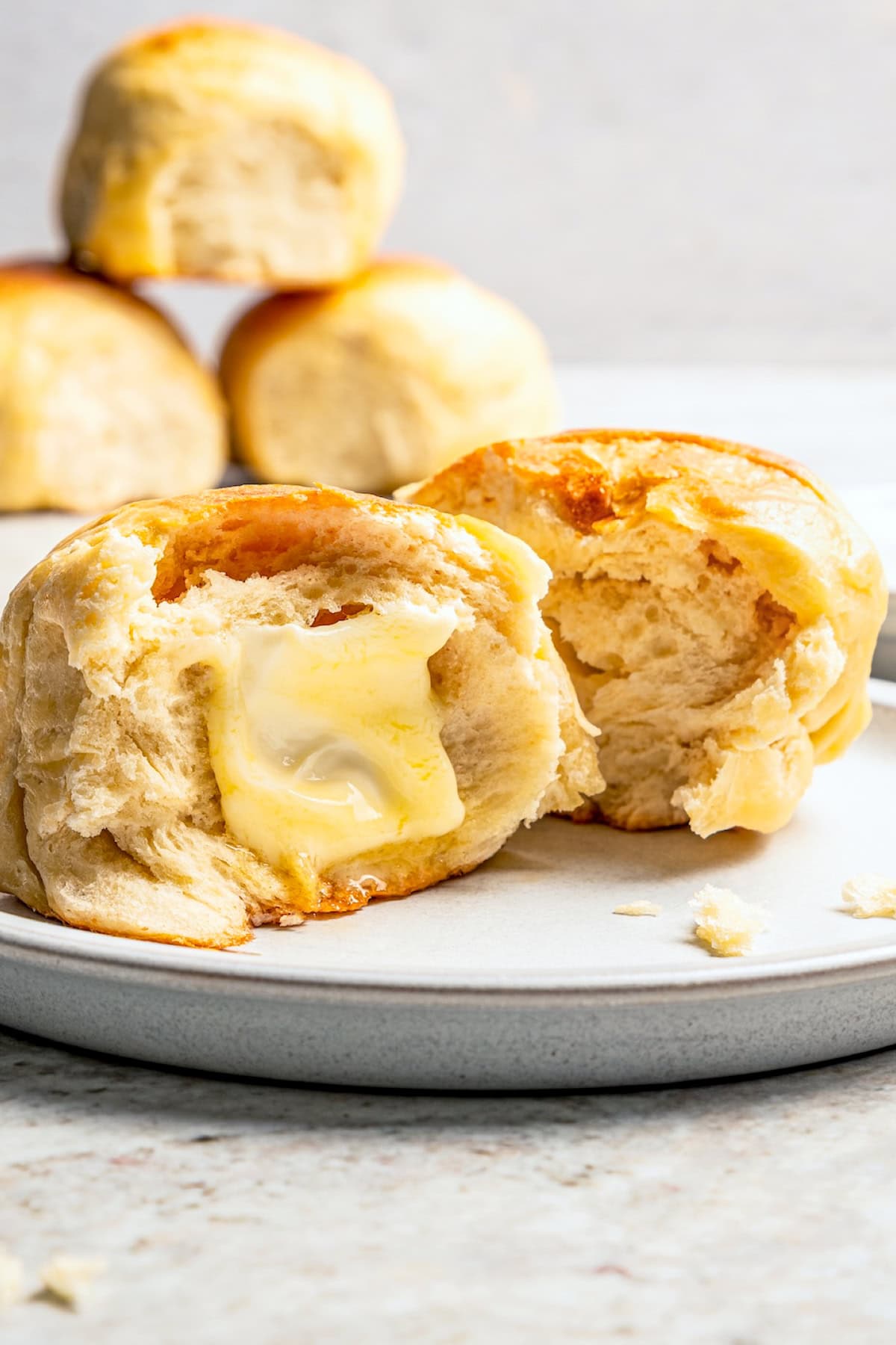 A Hawaiian roll broken in half, with one half buttered on a plate, and more rolls stacked in the background.