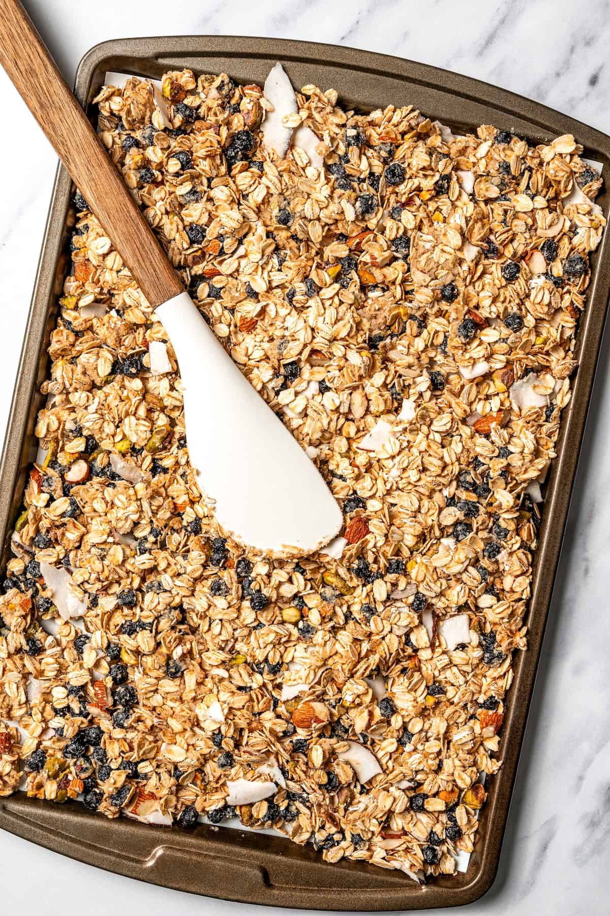 This homemade granola recipe with rolled oats, maple syrup, and brown sugar is sweet, crunchy, and you can make a big batch in the oven! A spatula resting on top of unbaked granola spread out on a baking sheet.