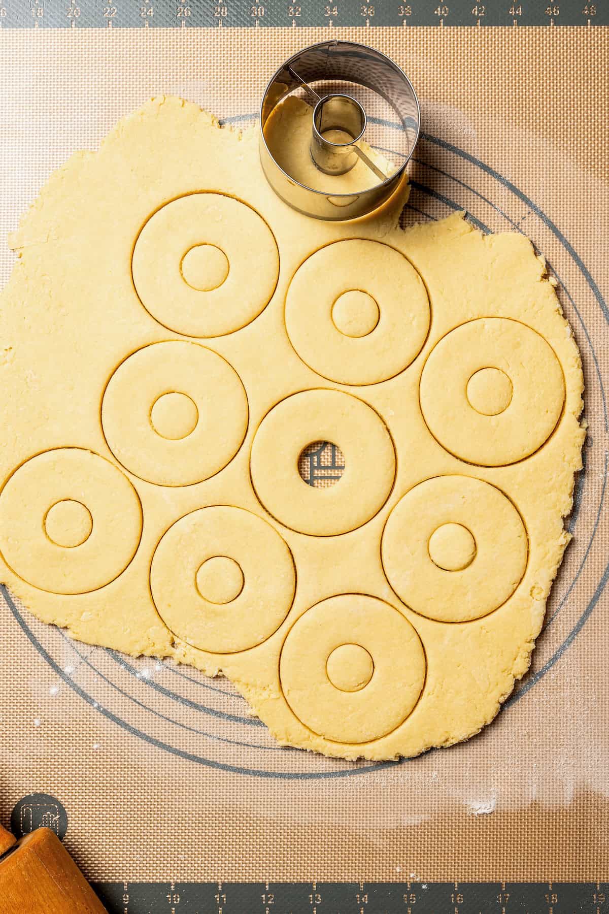 These old-fashioned sour cream donuts are soft and cakey beneath a golden crust coated in creamy vanilla glaze. No yeast or rising time! Donut rings being cut from a sheet of dough with a donut cutter.