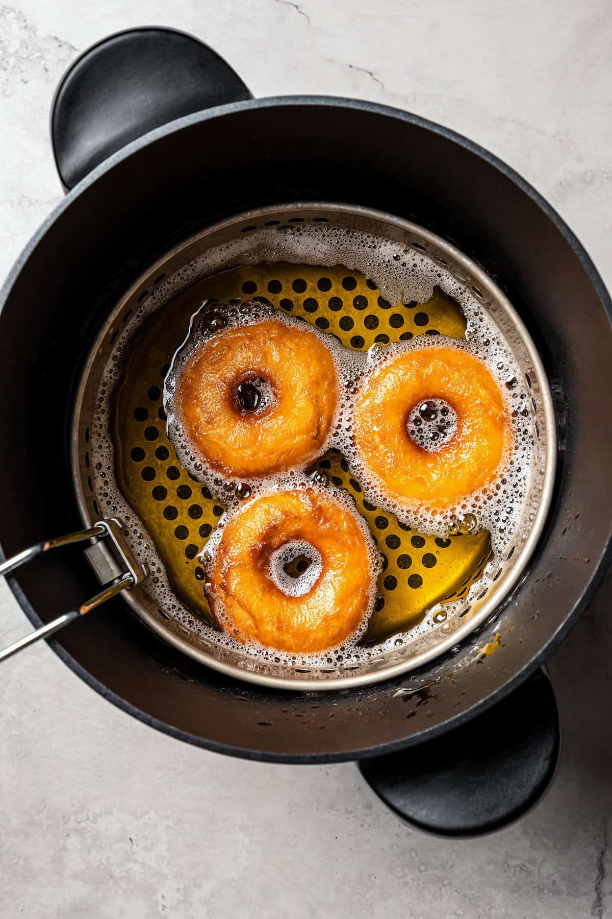 These old-fashioned sour cream donuts are soft and cakey beneath a golden crust coated in creamy vanilla glaze. No yeast or rising time! Three sour cream donuts inside the deep fryer.