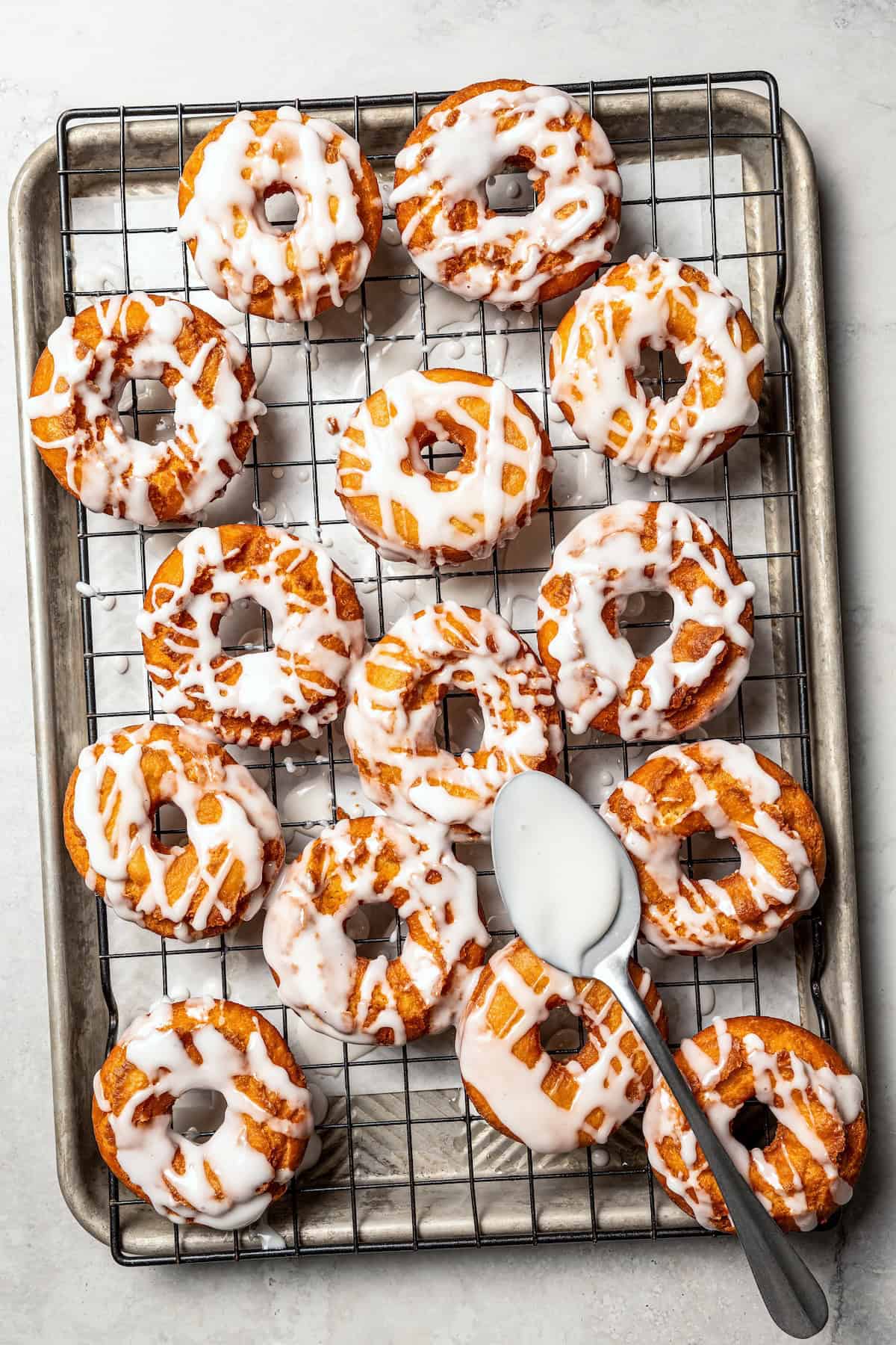 These old-fashioned sour cream donuts are soft and cakey beneath a golden crust coated in creamy vanilla glaze. No yeast or rising time! Overhead view of a spoon drizzling glaze over sour cream donuts on a wire rack.