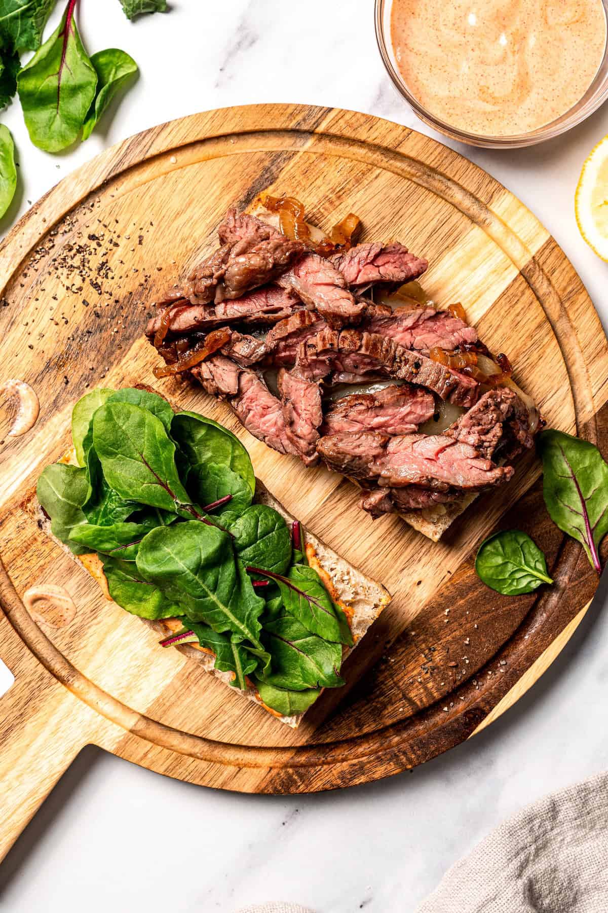 Overhead view of a partially assembled steak sandwich with steak strips on one half and spinach on the other.