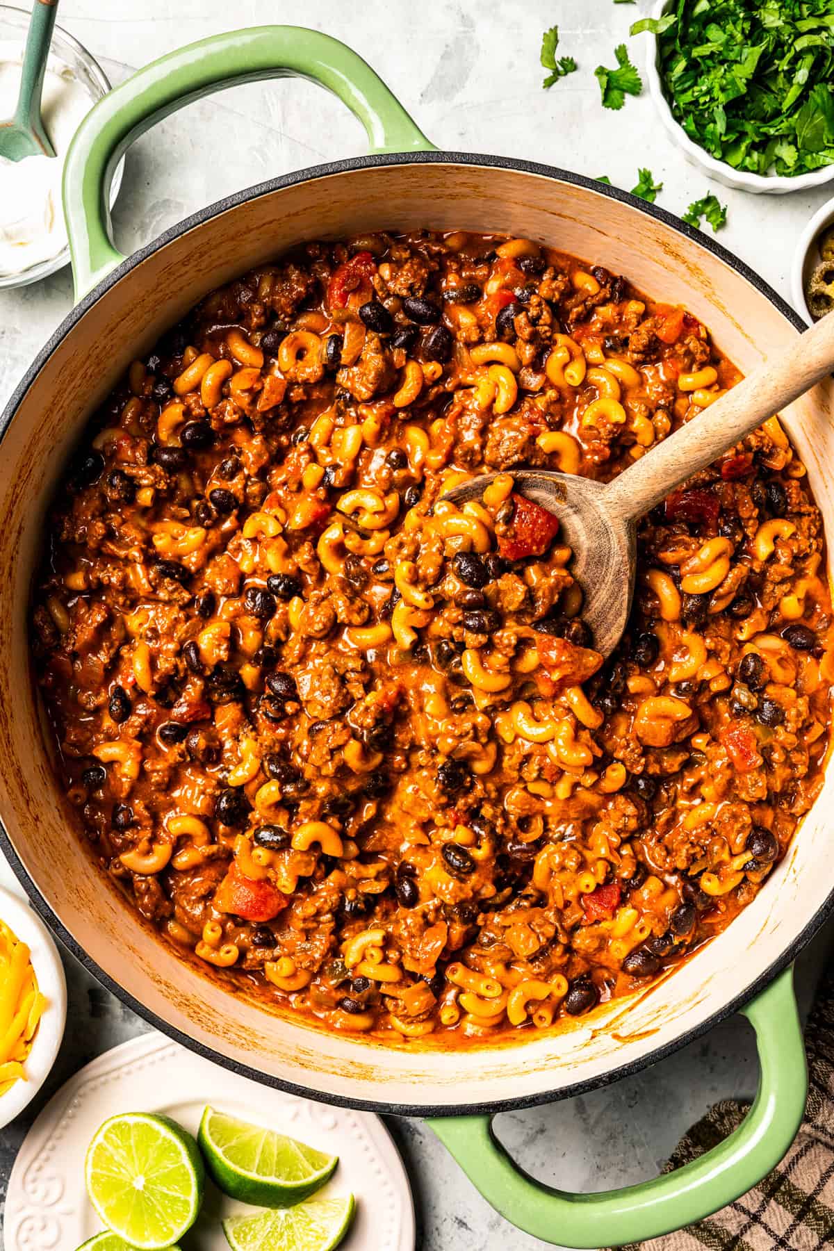 A pot of chili mac with a wooden spoon stirring through it.