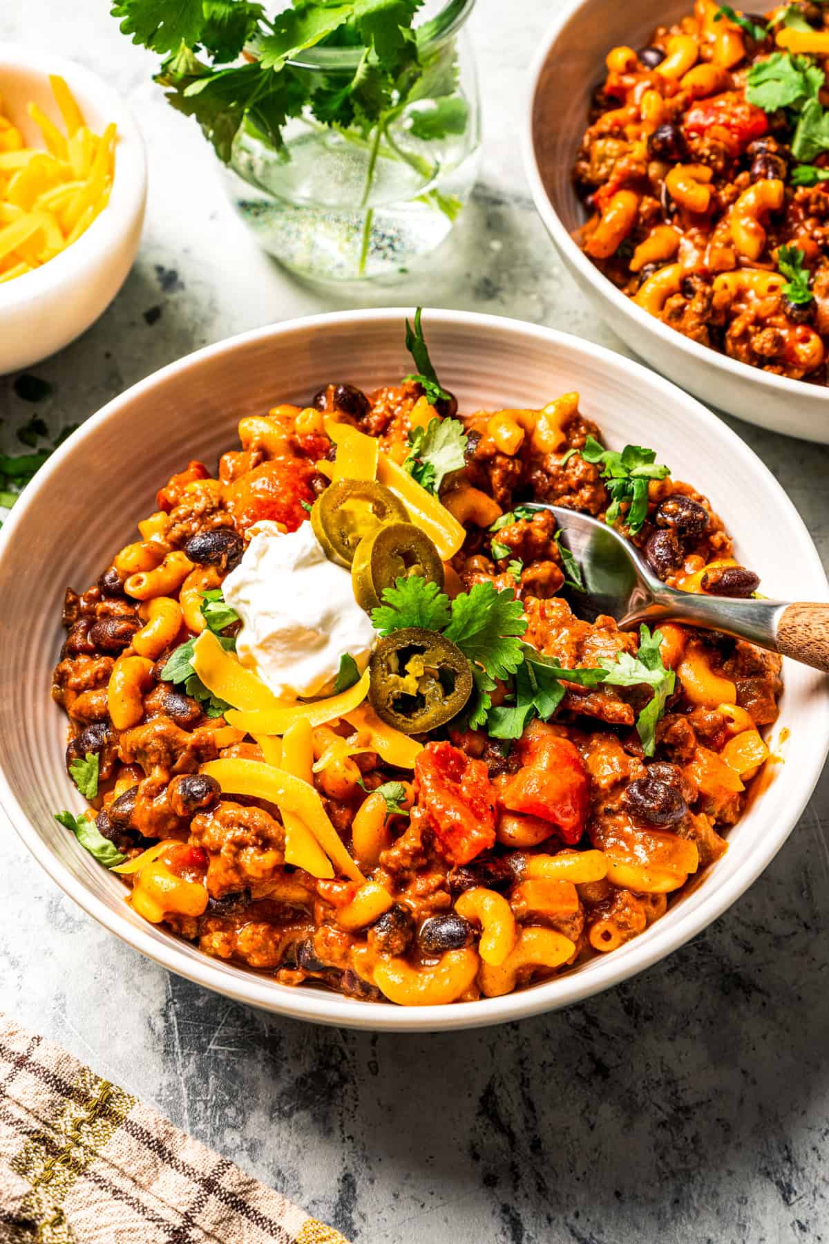 Bowls of chili mac garnished with sour cream, jalapeños, shredded cheese, and cilantro, surrounded by more toppings.