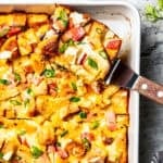 A slice of Eggs Benedict casserole is being lifted from the rest of the casserole in a baking dish.
