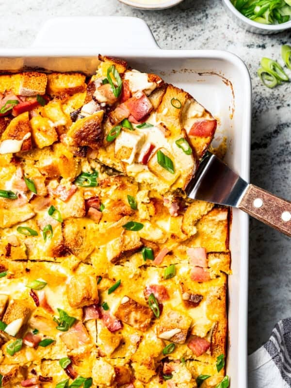 A slice of Eggs Benedict casserole is being lifted from the rest of the casserole in a baking dish.