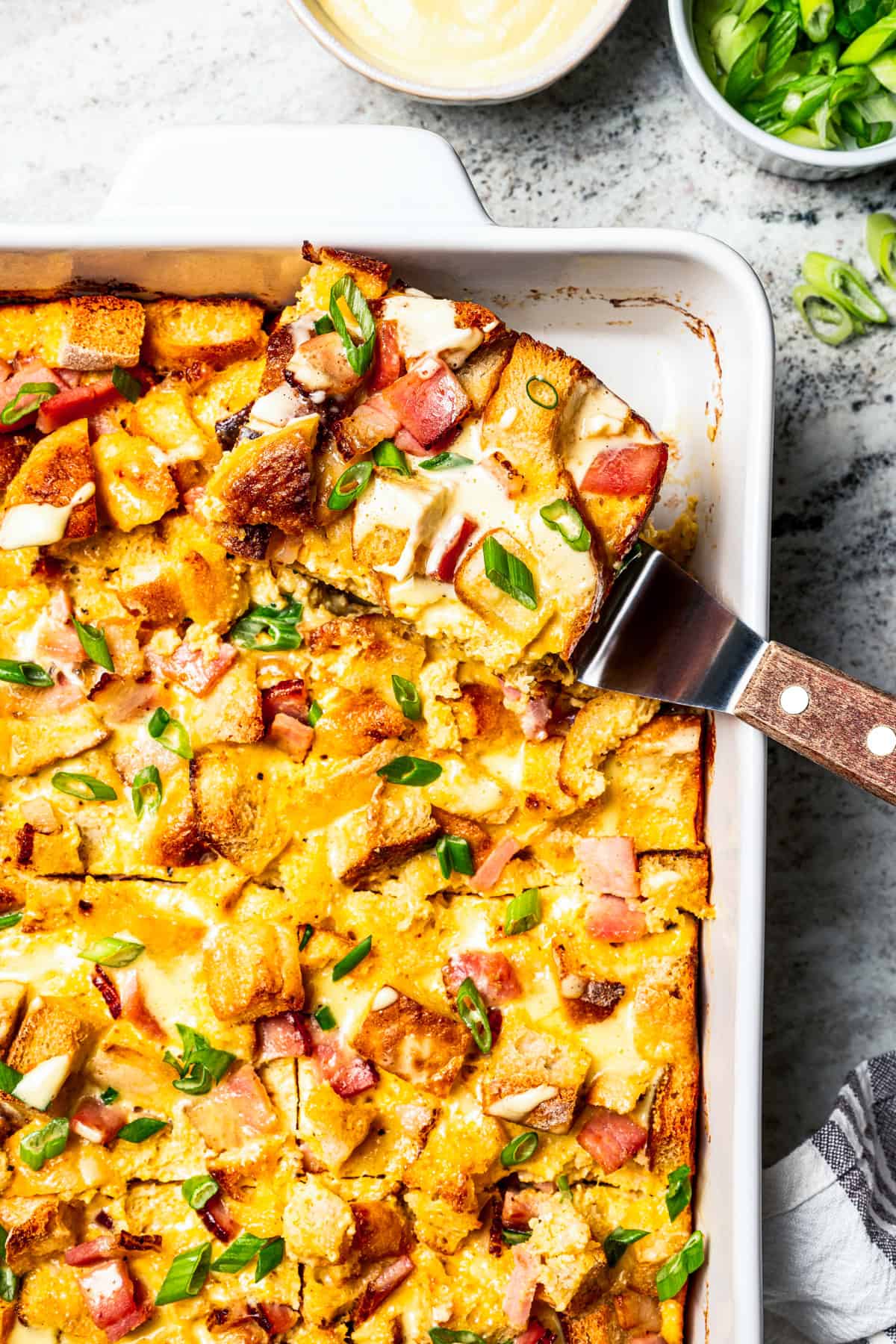 A slice of Eggs Benedict casserole is being lifted from the rest of the casserole in a baking dish.