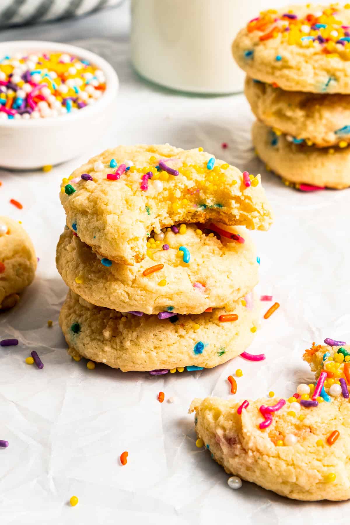 Stacks of funfetti cookies with a glass jug of milk and a bowl of sprinkles in the background.
