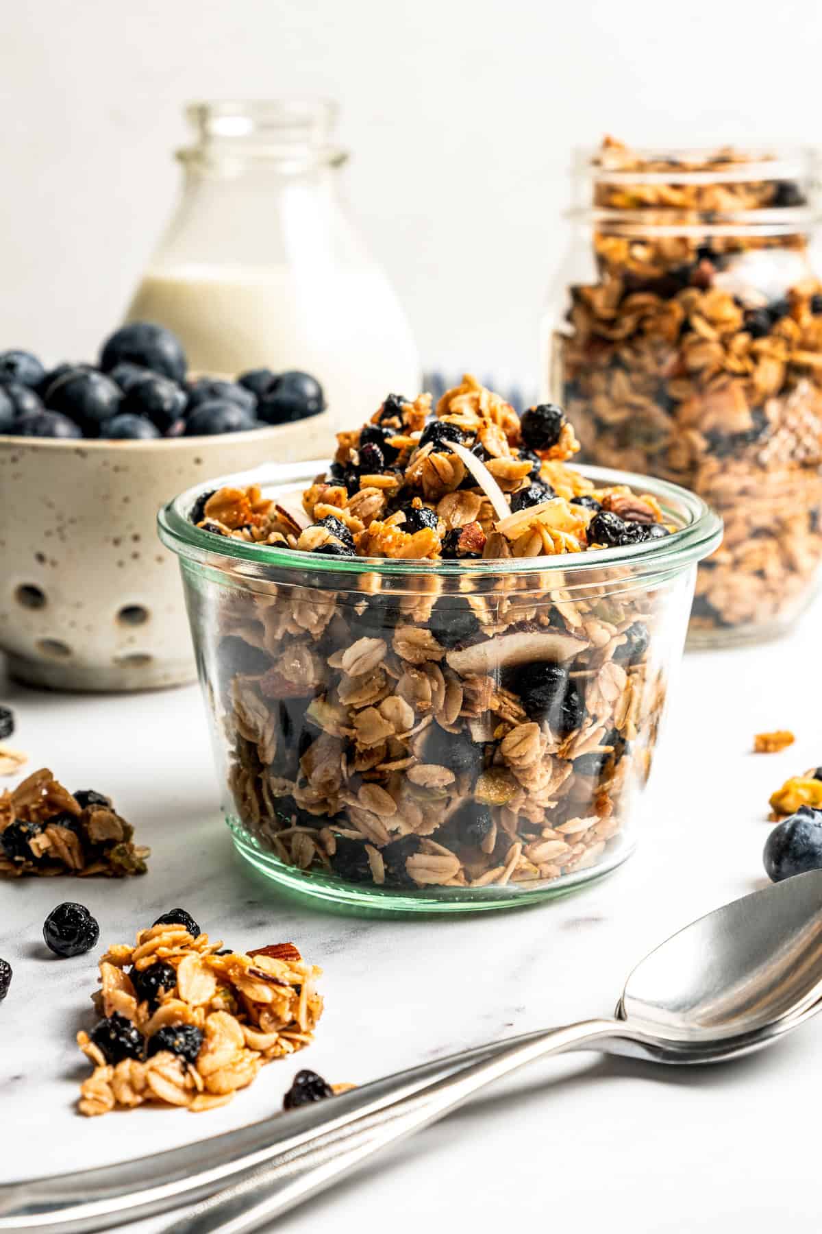 This homemade granola recipe with rolled oats, maple syrup, and brown sugar is sweet, crunchy, and you can make a big batch in the oven! Homemade granola in a small glass bowl, with a jug of milk, a jar of granola, and a bowl of blueberries in the background.
